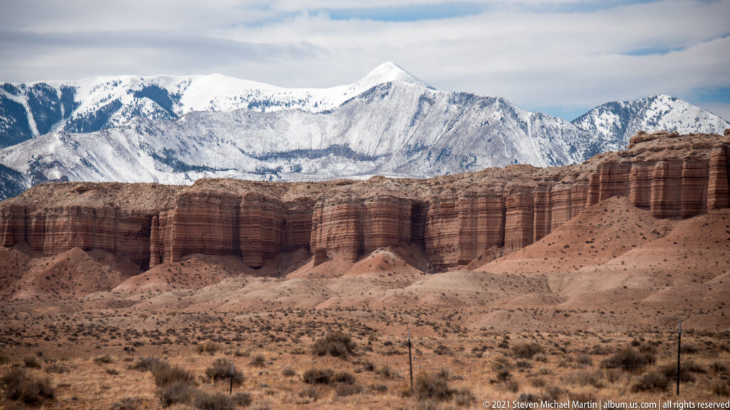 Snow-covered peaks rise behind a red-brown escarpment and scrub-covered desert slopes.