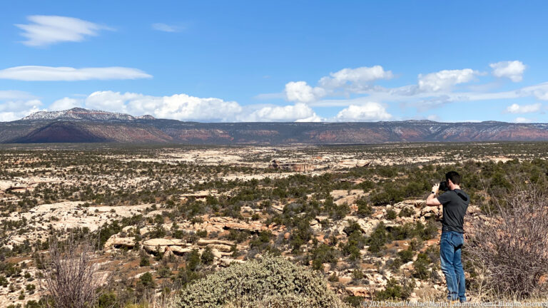 A visitor photographs a wide desert plateau from a rocky, shrub-covered overlook.