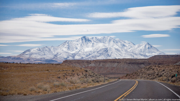 A two-lane highway curves through desert scrub toward a snow-covered mountain range.