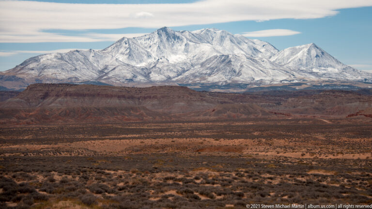 Snow-covered peaks rise above a rust-colored basin with sparse desert vegetation.
