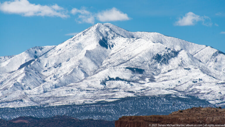 Snow-covered gullies and dark tree bands cover a steep mountain above a red-rock rim.