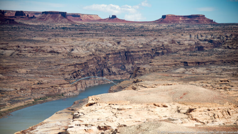 A steel arch bridge crosses a blue river far below a pale canyon overlook.