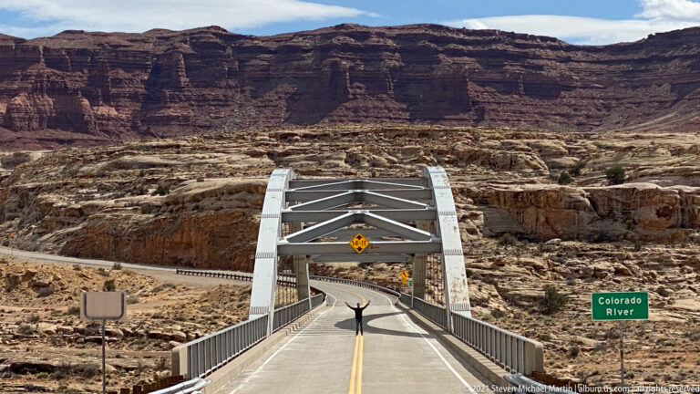 A visitor raises both arms beneath a steel bridge arch, with canyon cliffs and a Colorado River sign behind.