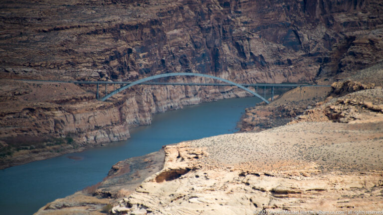 A steel arch bridge spans blue water below a pale ledge and dark canyon walls.
