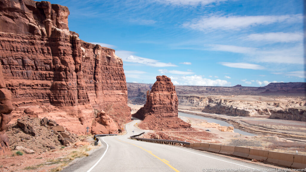 A paved road curves around a red sandstone tower beside a high cliff.