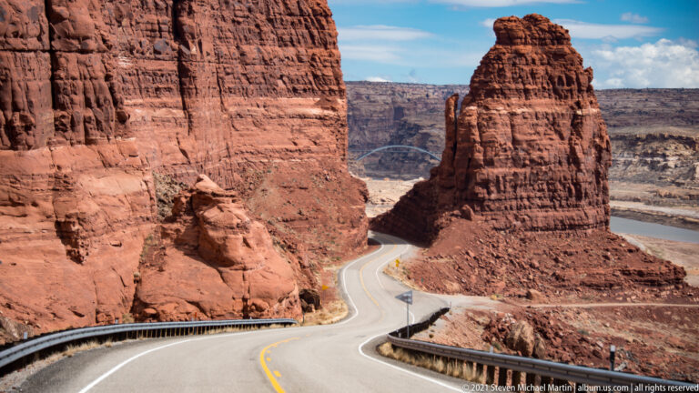 A curving road passes between red cliffs and a tall sandstone tower above a distant canyon floor.