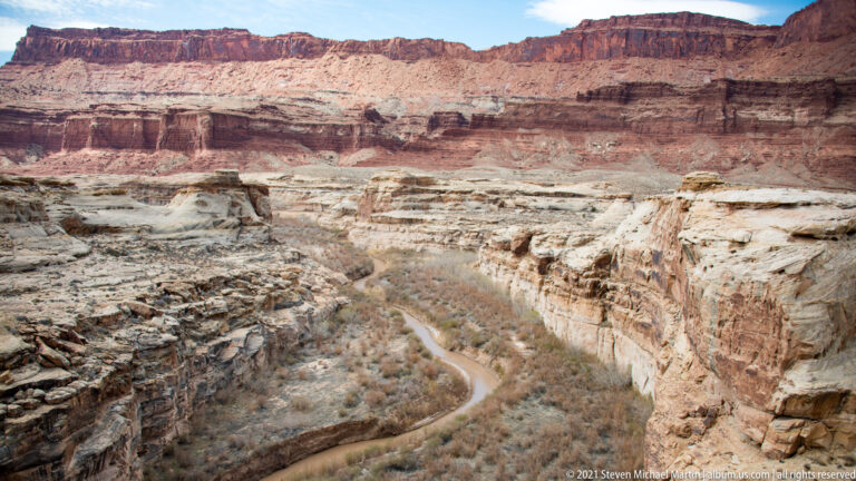 A narrow stream curves through a dry canyon lined with pale rock shelves and red walls.
