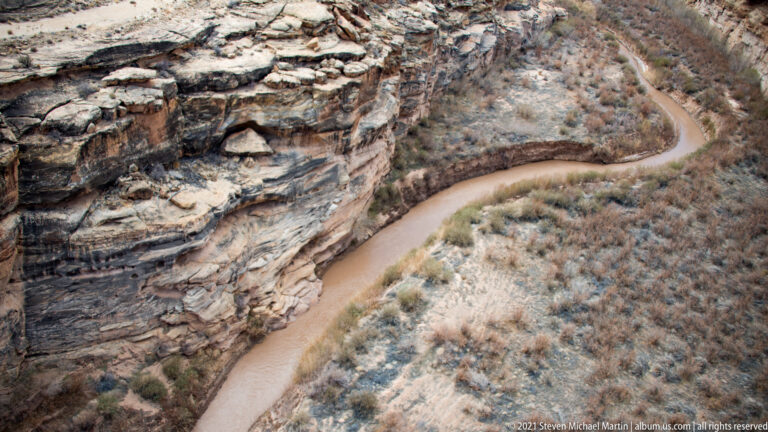 A muddy stream curves beside fractured, layered canyon rock and leafless trees.