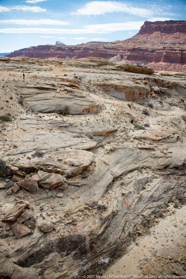 A distant visitor crosses a broad pale rock bench below red mesas.