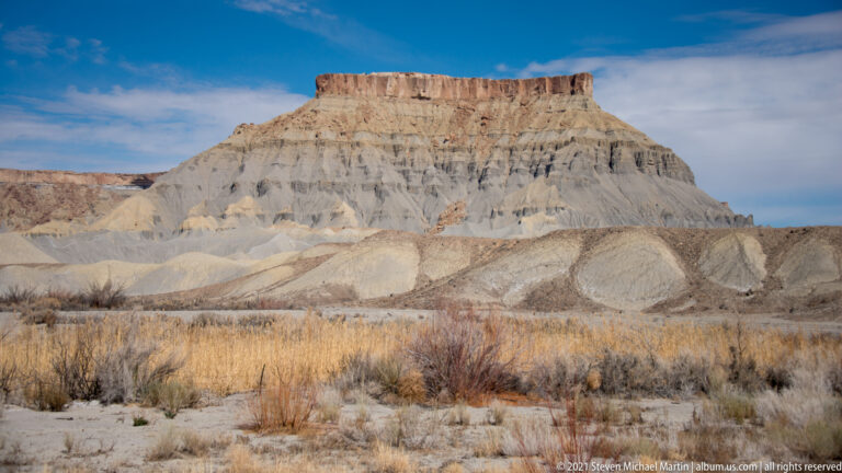 A flat-topped pale butte rises above dry grasses and low shrubs beneath a blue sky.