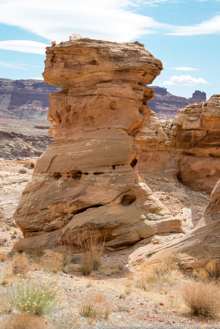A layered sandstone pedestal with a broad cap stands among dry grasses, with distant mesas behind it.