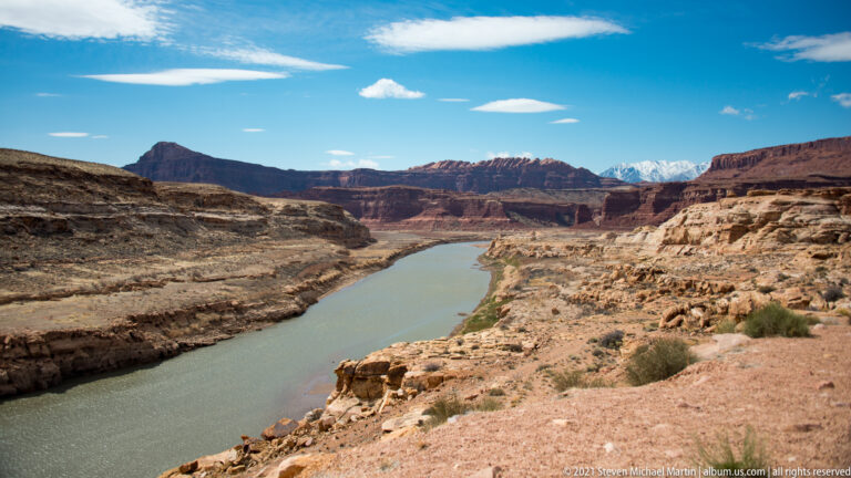 The green Colorado River bends between pale rocky banks and layered red canyon walls beneath a blue sky.