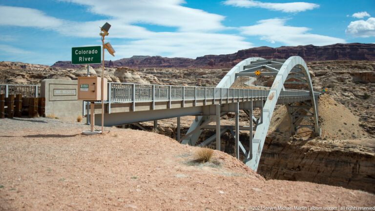 A steel arch bridge and fenced walkway cross a rocky gorge beside a green Colorado River sign.