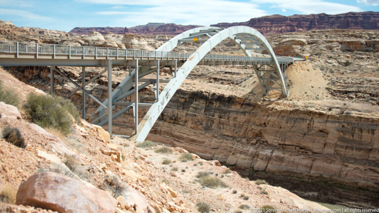 A pale steel arch bridge spans a deep rocky gorge, seen from the side above the canyon slope.