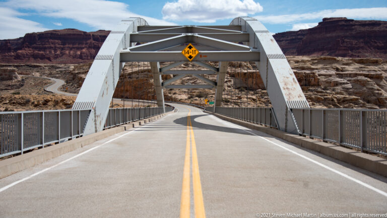 A two-lane roadway passes through white steel bridge arches beneath a yellow 14-foot-11-inch clearance sign.