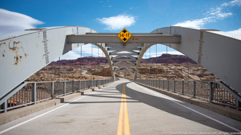 The bridge roadway and railings lead beneath a white steel arch toward the canyon, under a yellow clearance sign.