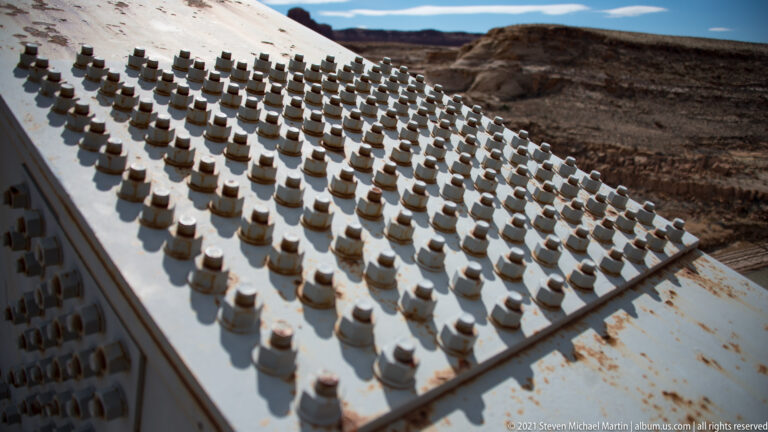Rows of bolt heads and light rust marks cover a slanted pale steel bridge plate above the canyon.