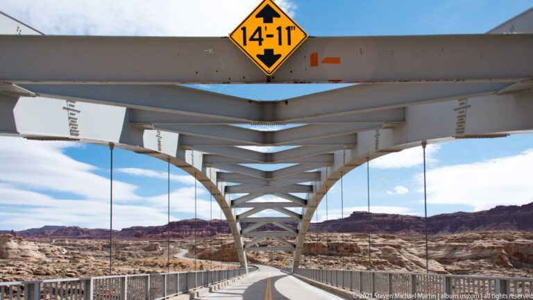 Steel cross-braces and a curved arch frame the roadway and canyon beyond from beneath the bridge.