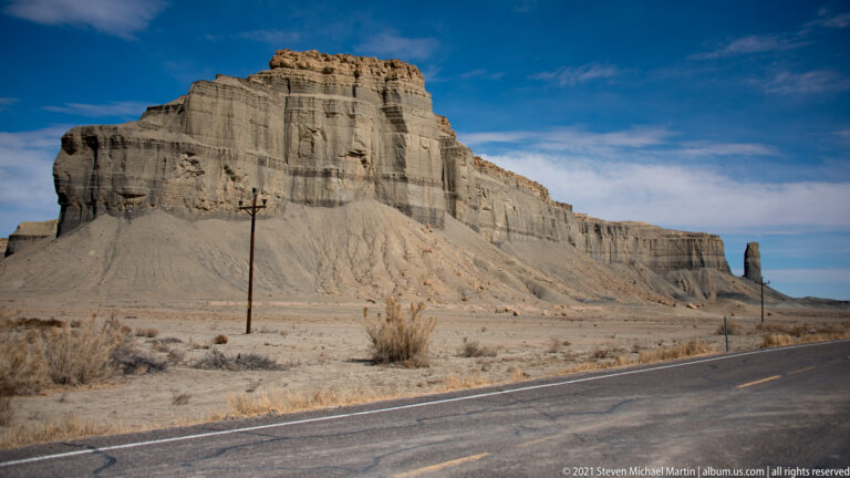 A pale, layered sandstone cliff rises above a highway, with a utility pole in the foreground.