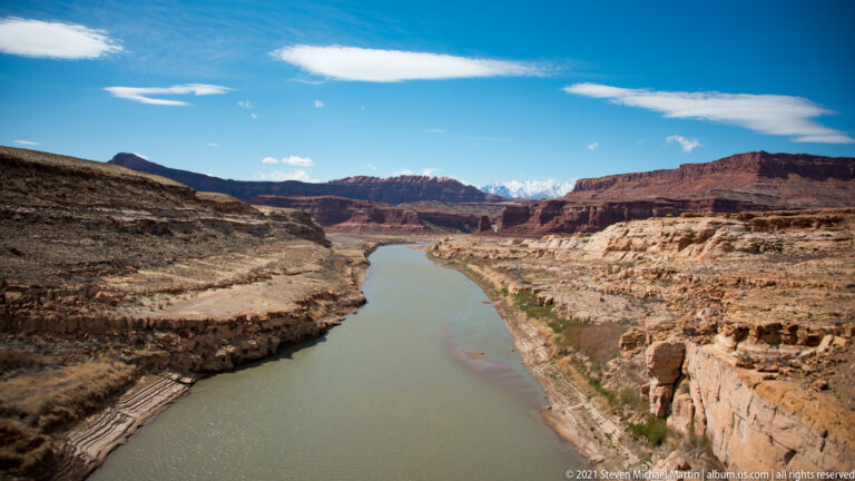 The Colorado River curves through a wide canyon bordered by layered cliffs and pale rocky benches.