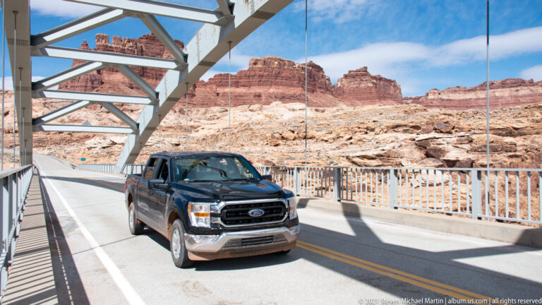 A dark pickup truck drives across a steel arch bridge, with canyon cliffs visible beyond the railings.