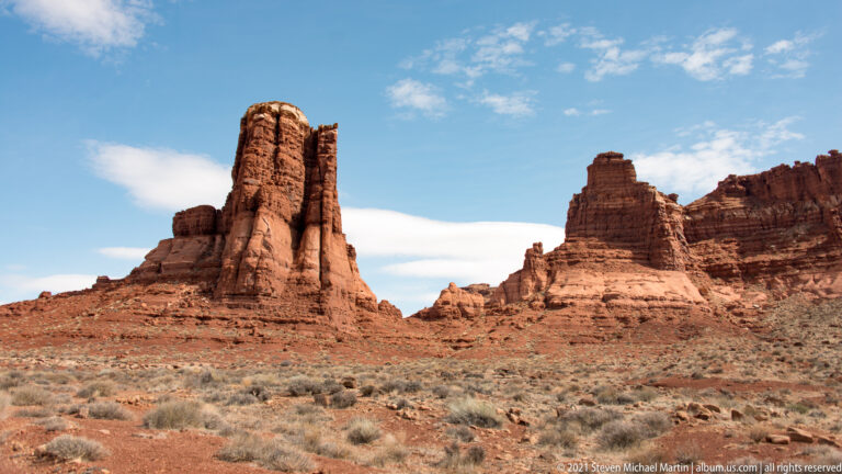Red sandstone towers rise above sparse desert shrubs beneath a blue sky with thin clouds.