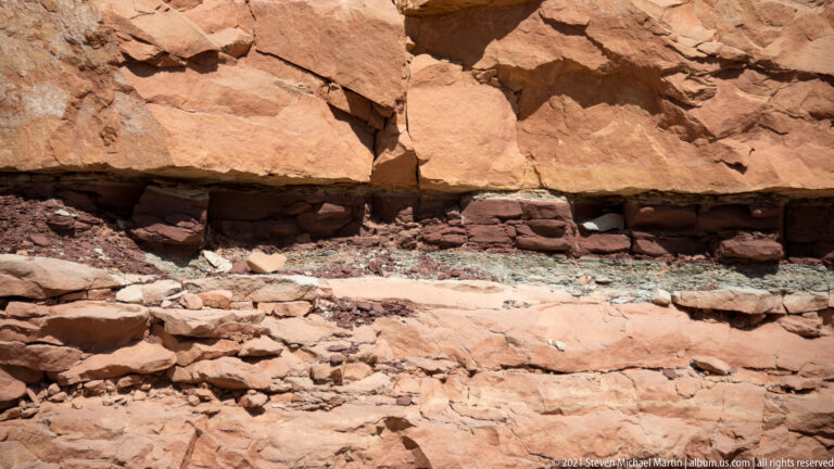 A broad orange sandstone ledge shelters a dark recess above fractured rocks and smaller stones.