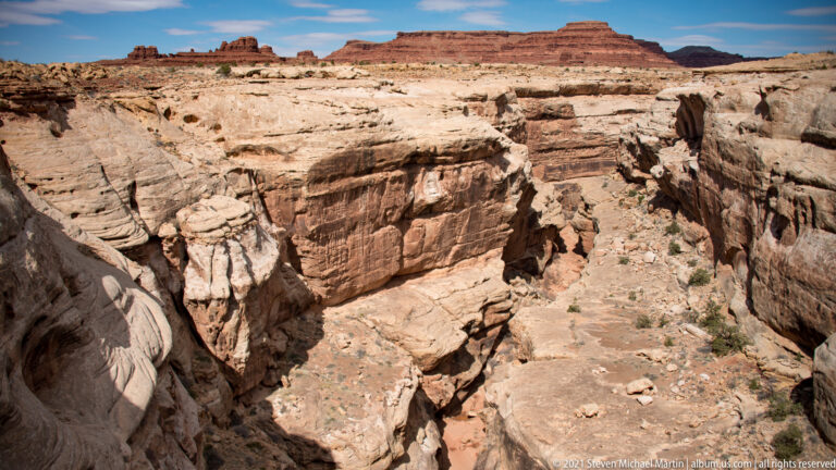 A deep sandstone gorge with stepped walls descends from a rocky rim toward a distant mesa.
