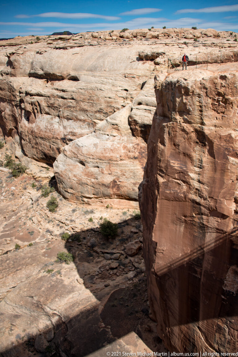 A small hiker stands on a pale sandstone bench above a dark cleft and rounded foreground slab.