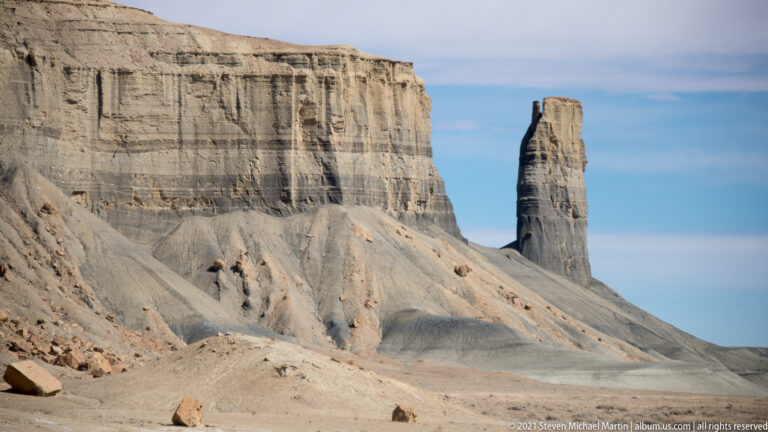 A narrow sandstone pillar stands beside a broad pale cliff above an eroded desert slope.