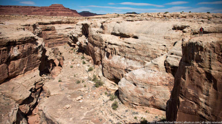A narrow deep canyon splits pale sandstone walls, with a small hiker standing near the upper rim.