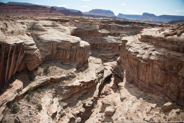 A sunlit branching canyon stretches from a rocky rim toward a low mesa in the distance.