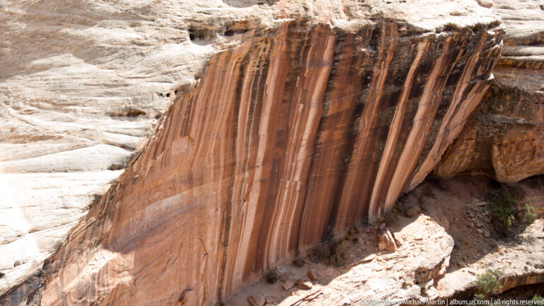 Vertical rust-colored bands stripe a pale sandstone wall above a broken rocky ledge.