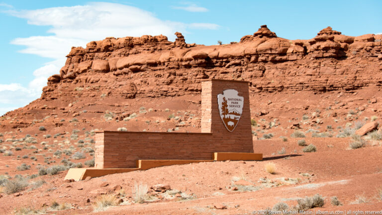 A stone monument with a National Park Service arrowhead stands before a red sandstone hillside.