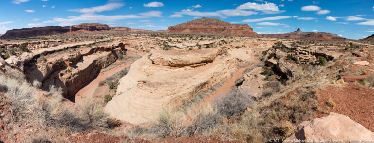 A broad sandstone ravine crosses the foreground beneath a flat-topped mesa and scattered clouds.