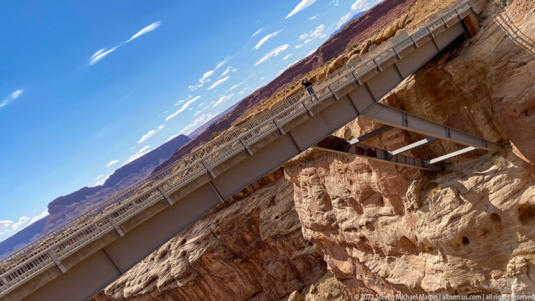 Steel bridge beams cross a red-rock gorge, viewed from below at a diagonal angle.