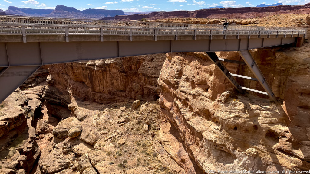 A steel bridge spans an eroded gorge, with its supports descending toward a rocky canyon floor.