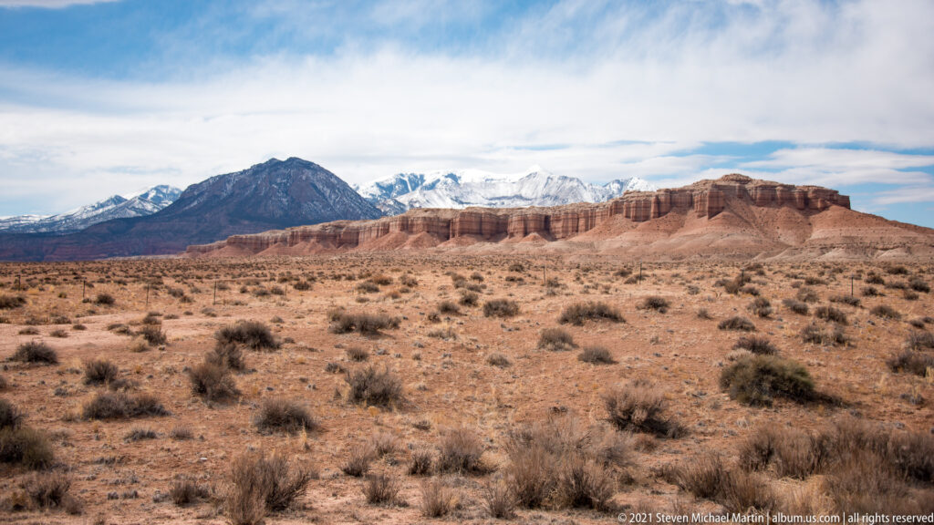 Red and cream mesas cross a desert plateau below a distant snow-dusted mountain.