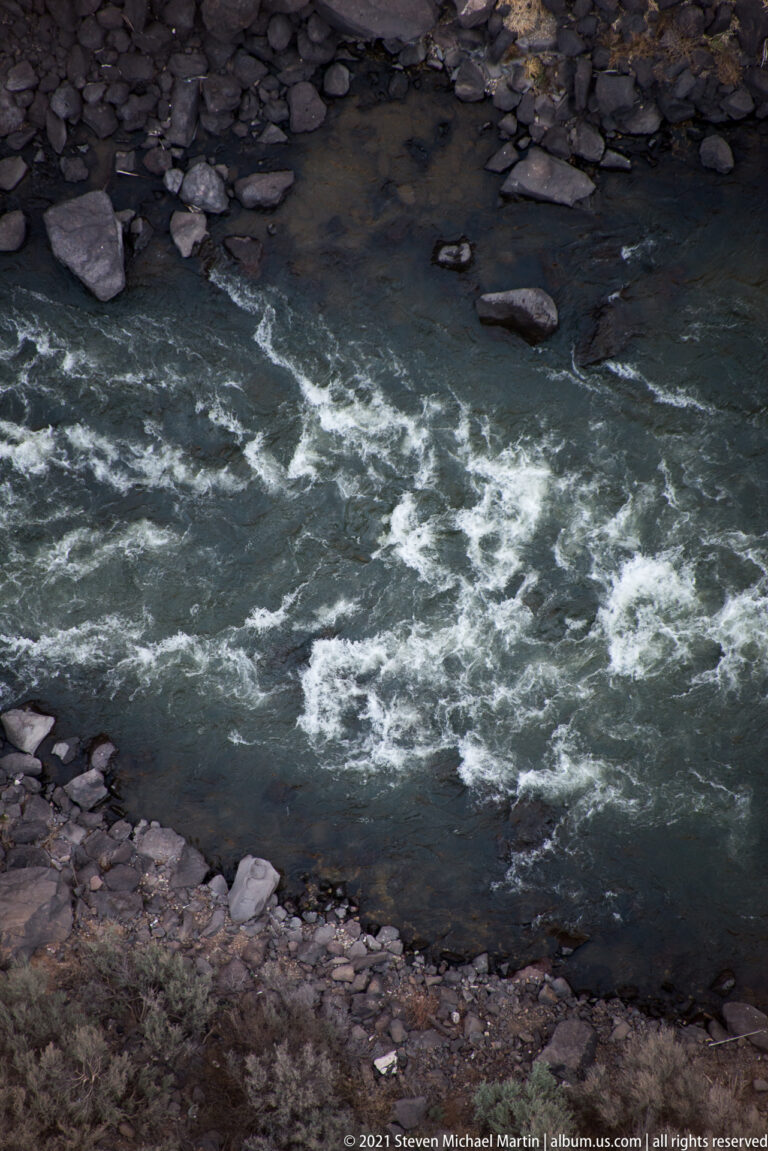 Gray-green water breaks into white rapids around dark rocks beside a narrow bank.