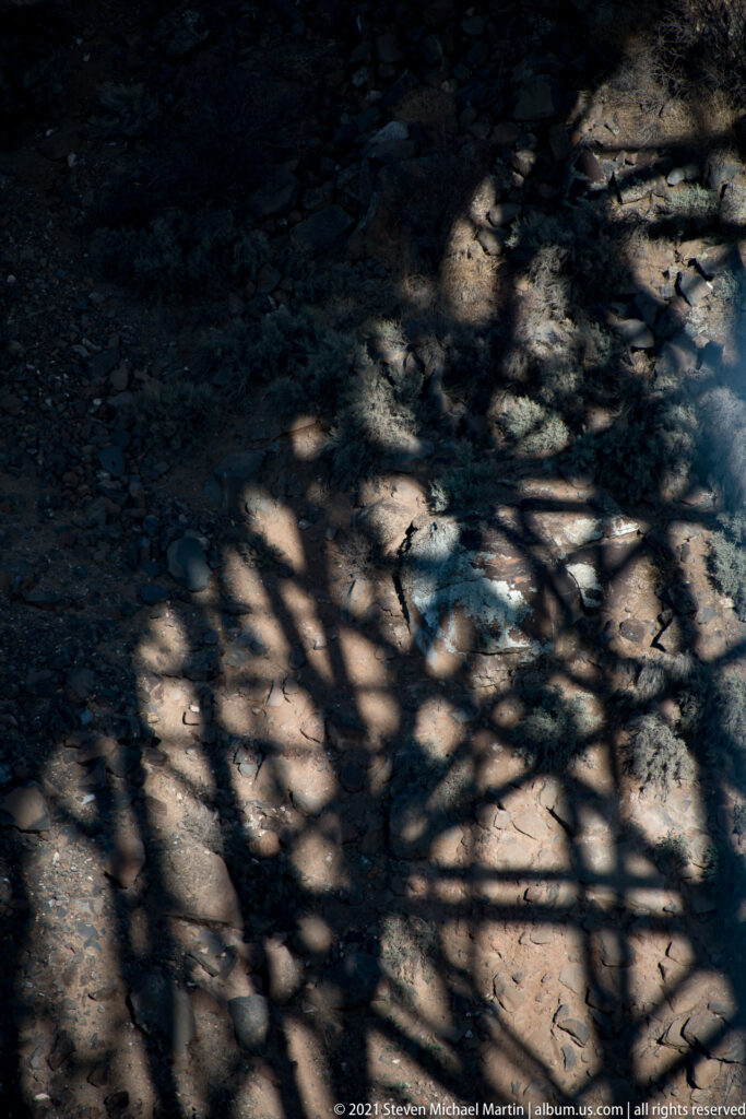 A geometric bridge shadow falls across shallow water and rounded rocks in the riverbed.
