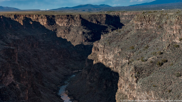 A narrow river runs through a deep shadowed gorge between steep rocky walls and distant mountains.