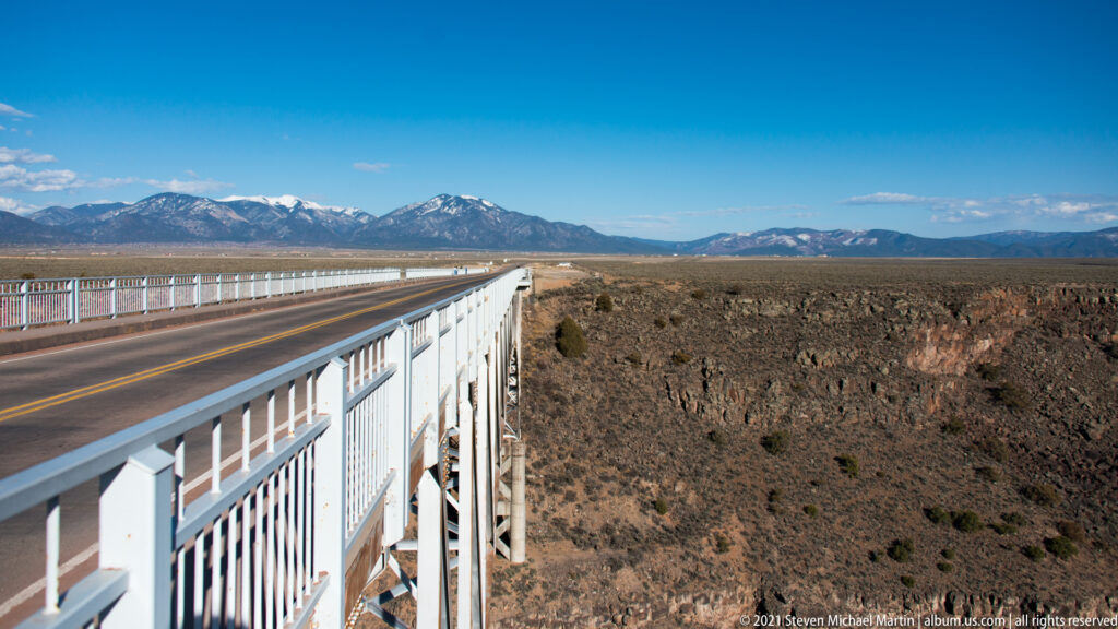 A road and white railing cross the gorge toward distant mountains, with bridge supports below.