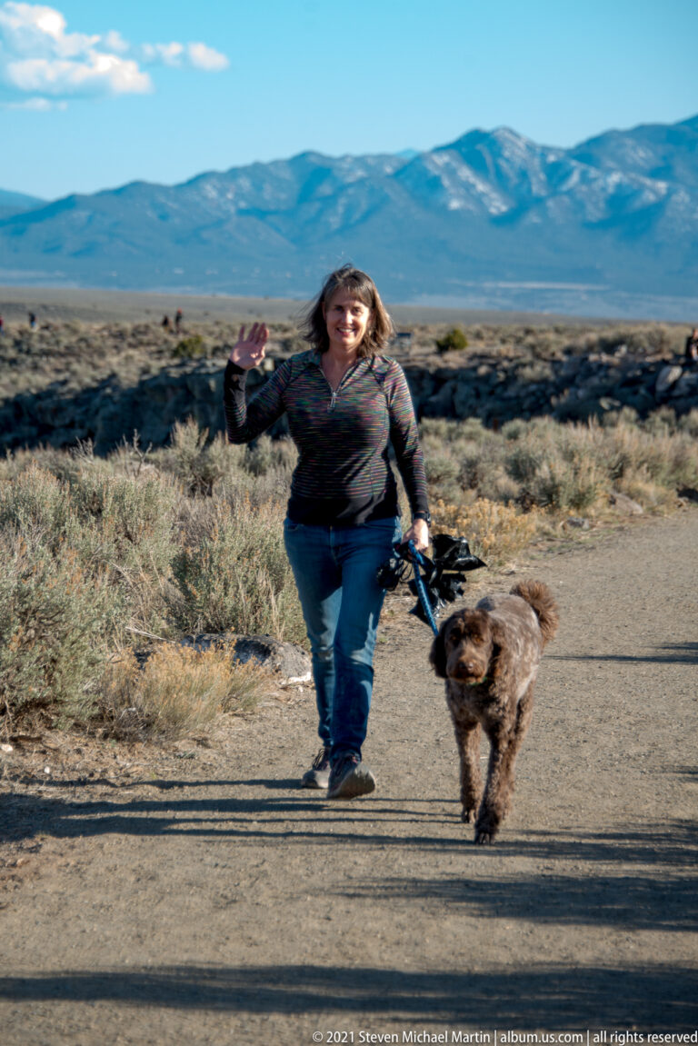 A woman raises one hand while walking a leashed brown dog on a paved high-desert path.