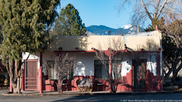 A low stucco building with red trim and a shaded porch stands beneath trees beside a road.
