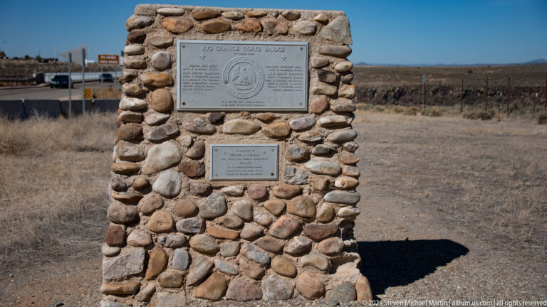 A stone pillar with two metal plaques stands in an open scrubland landscape.