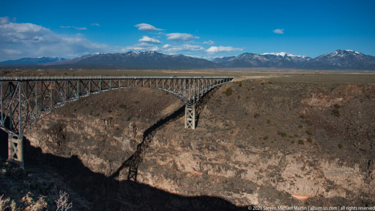 A steel truss bridge crosses a rocky gorge, casting a broad shadow on the canyon below.