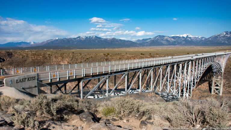 A bridge railing marked “Last Kiss” recedes across the gorge toward distant mountains.