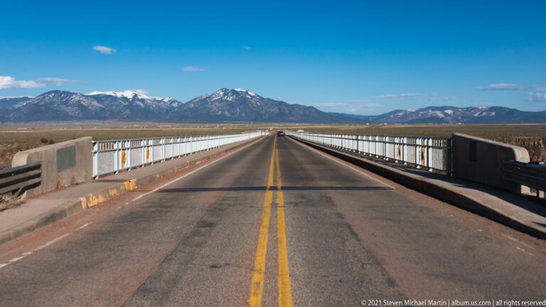 A two-lane road crosses a bridge toward distant mountains, framed by white railings.