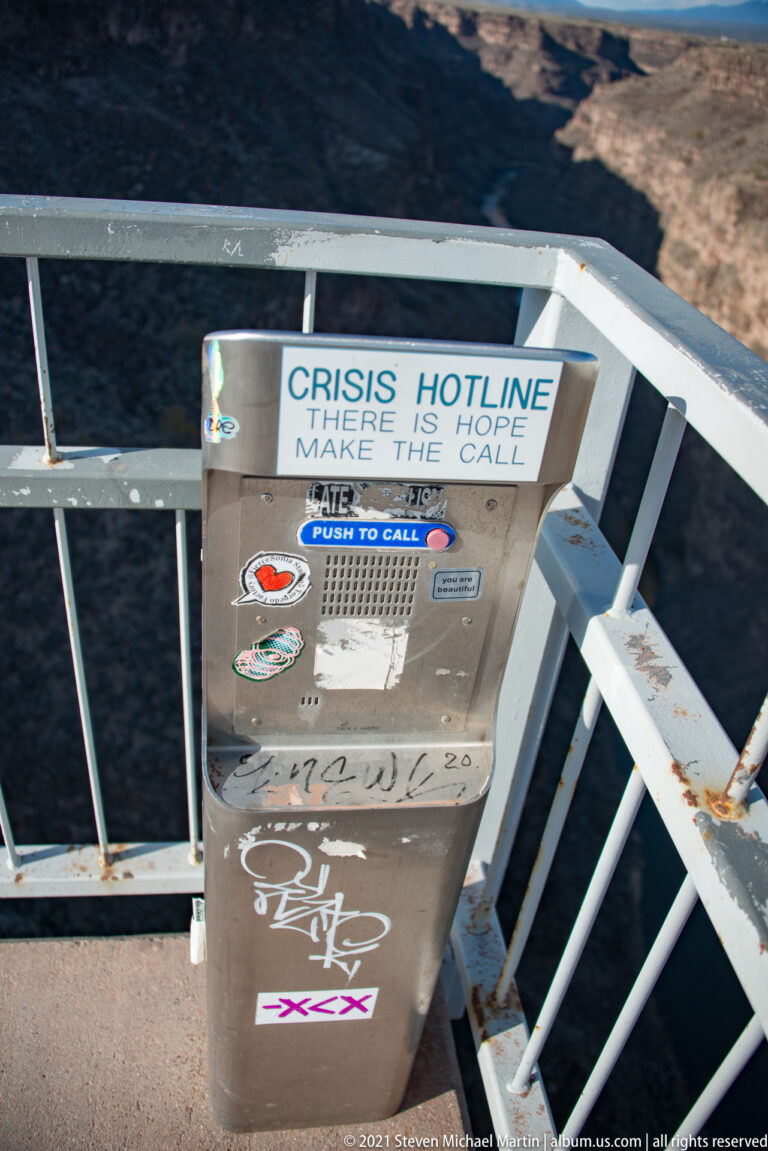 A metal crisis hotline call box covered with stickers is attached to a white bridge railing.