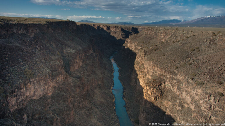 A broad canyon rim overlooks the shadowed river channel and distant mountains.